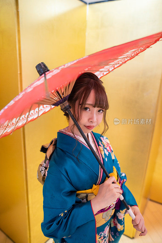 身着“furisode”和服的年轻女子手持纸伞，站在金色的“Byobu”折叠屏风前，参加“Seijin Shiki”成人礼